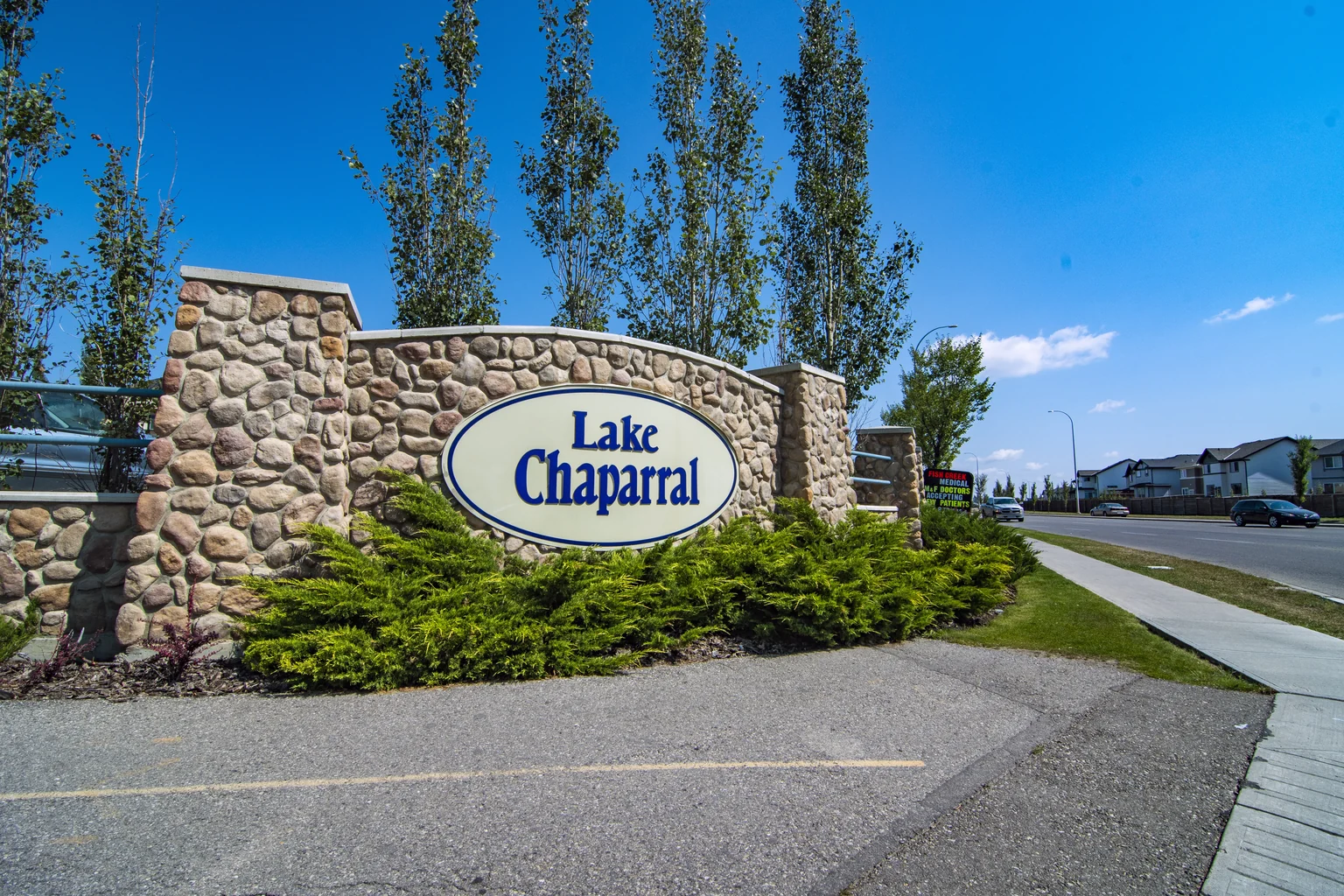 Lake Chaparral entrance sign set in a curved stone wall with shrubs and tall trees beside the sidewalk