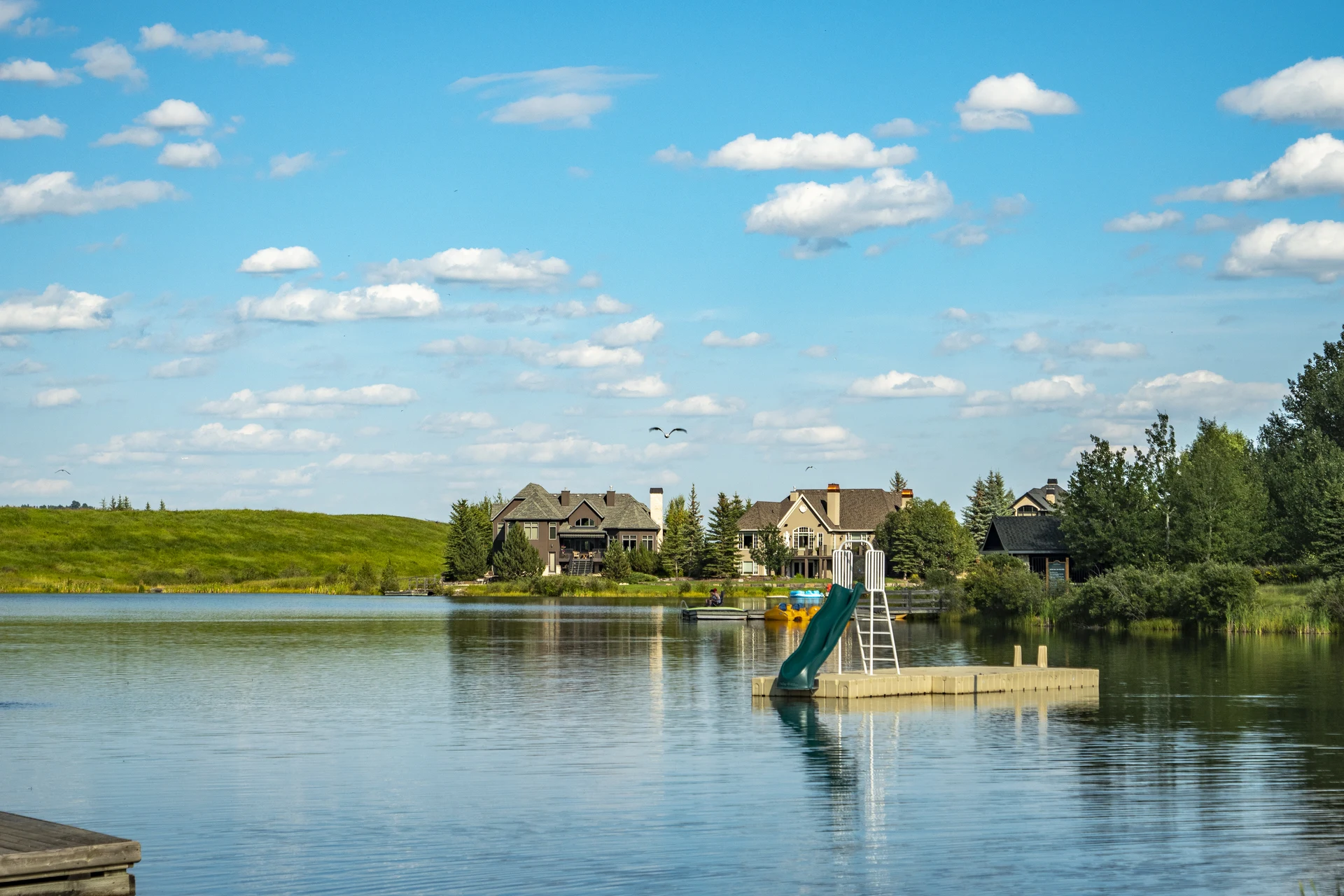 Elbow Valley lake with a green slide on a floating platform, shoreline pavilion and homes