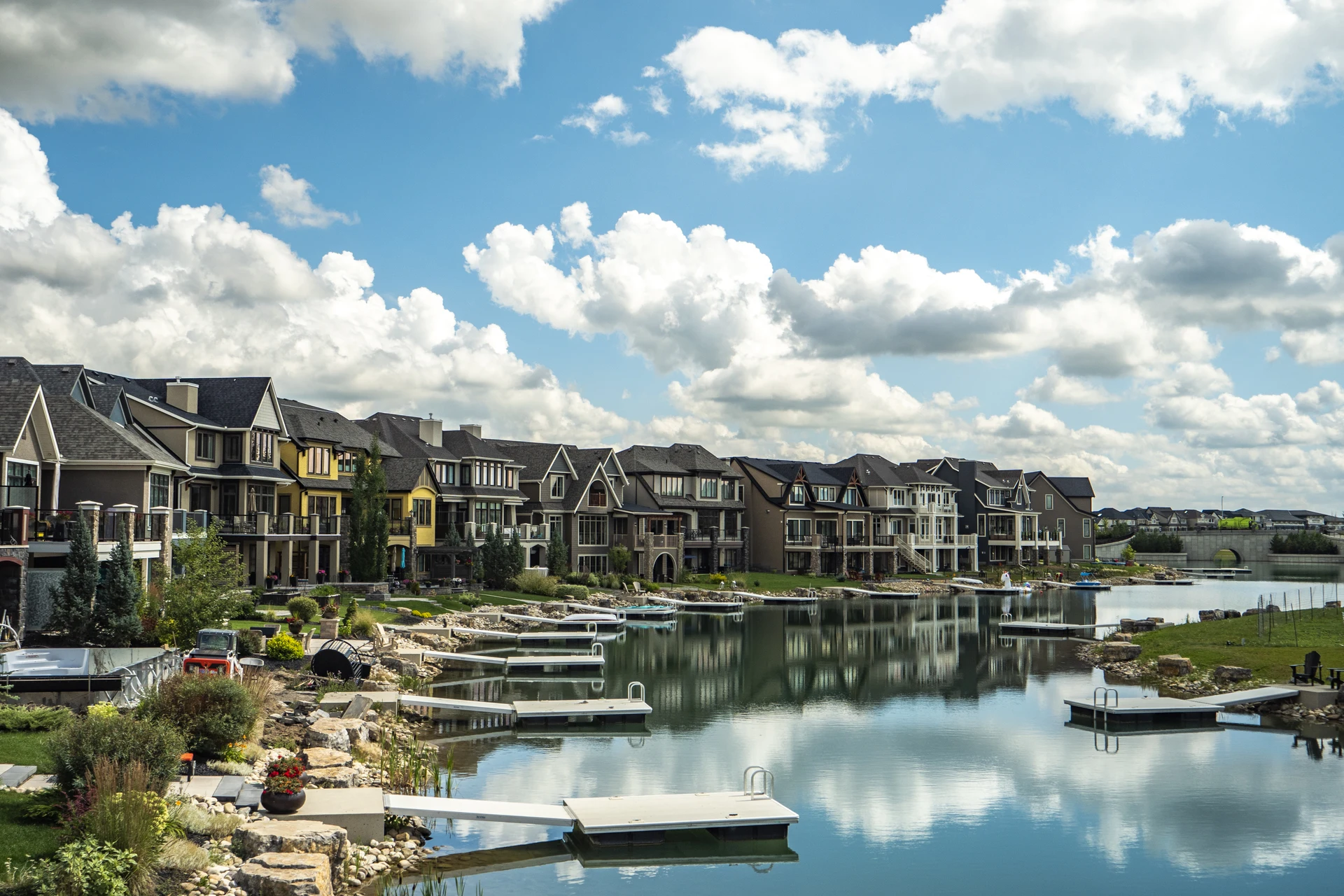 Mahogany waterfront homes and private docks beside reflective lake water, with an arched bridge in the distance