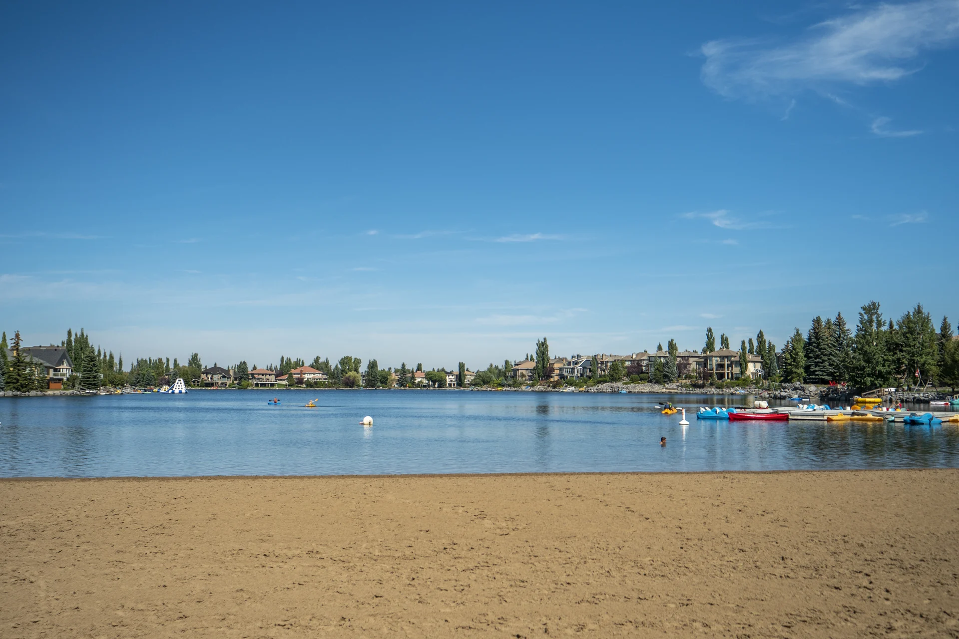 McKenzie Lake seen across a sandy beach, with waterfront homes and trees beyond the water and small boats along the right shoreline